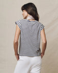 A woman with dark hair faces away, wearing The Great Inc.'s The Peak Shoulder Tee in black and white stripes, paired with white pants and a patterned neck scarf, set against a neutral background.
