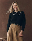 A woman with long blonde hair smiles, wearing The Great Inc.'s The Crochet Collar Pullover, a black sweater with a crochet collar, paired with a tan pleated skirt and decorative belt, standing against a brown textured backdrop.