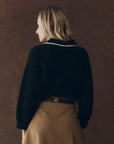 A woman with blonde hair wears The Great Inc.'s The Crochet Collar Pullover, a black sweater with a crochet collar, and a tan skirt as she stands facing away from the camera against a plain brown background.