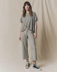 A woman with wavy brown hair stands against a beige backdrop, wearing The Great Inc. Cropped Sweat Pant in grey cotton terry with a matching t-shirt and black Converse high-tops, looking relaxed.