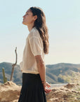 A woman with long hair stands outdoors in profile, wearing The Crochet Polo by The Great Inc. in cream mercerized cotton and a black pleated skirt. She faces the sun with her eyes closed, mountains and desert plants behind her.