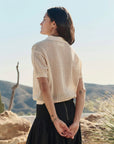 A person with long dark hair stands outdoors, hands clasped behind their back, wearing The Crochet Polo by The Great Inc. in cream, paired with a black skirt, with desert hills and cacti in the background.