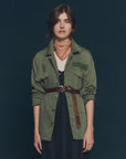 A woman with loosely styled brown hair and a neutral expression stands against a dark background, wearing The Vintage Army Jacket by The Great Inc. in cotton slub twill, belted over a black dress with a patterned red neck scarf.