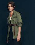 Against a dark background, a person with dark hair wears The Great Inc.'s The Vintage Army Jacket belted at the waist over a black dress, accessorized with a light patterned bandana around their neck.