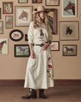 A woman in a cowboy hat, western graphic shirt, long floral embroidered skirt, and cowboy boots stands before a beige wall with framed horse art, her look completed by The Great Inc. Leather Concho Belt for true Western style.