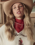 A woman with long, wavy blonde hair wears a tan cowboy hat, The Stetson Western Bandana by The Great Inc. around her neck, and a cream sweater with a western-themed graphic as she gazes upward in soft light.