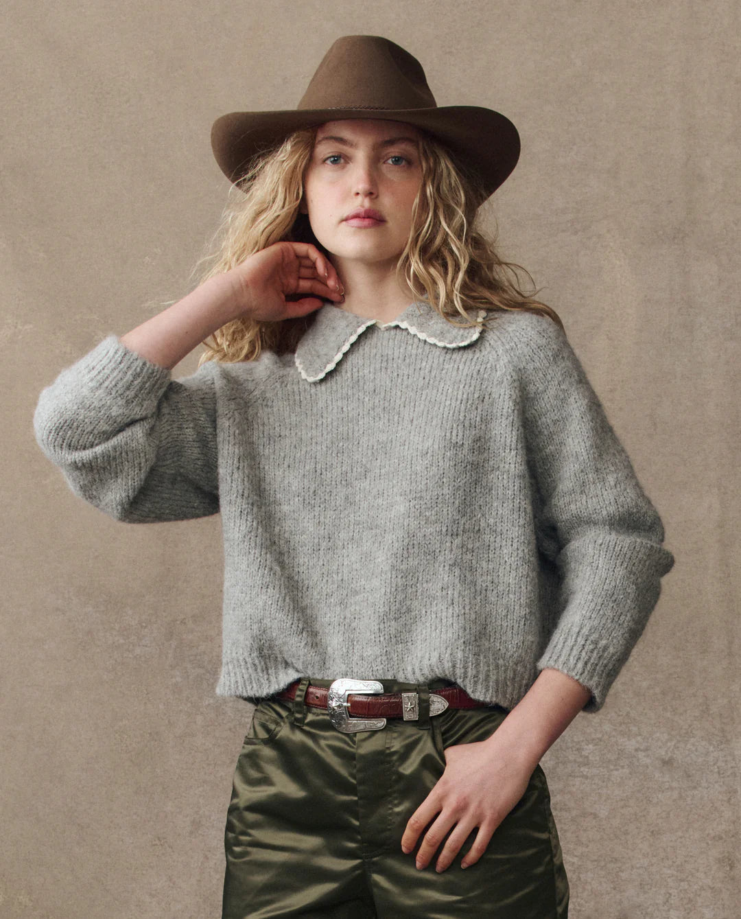A young person with long, wavy blonde hair wears The Great Inc.'s Crochet Collar Pullover and a brown wide-brimmed hat, paired with green satin pants and a silver-buckled belt, standing against a neutral background.