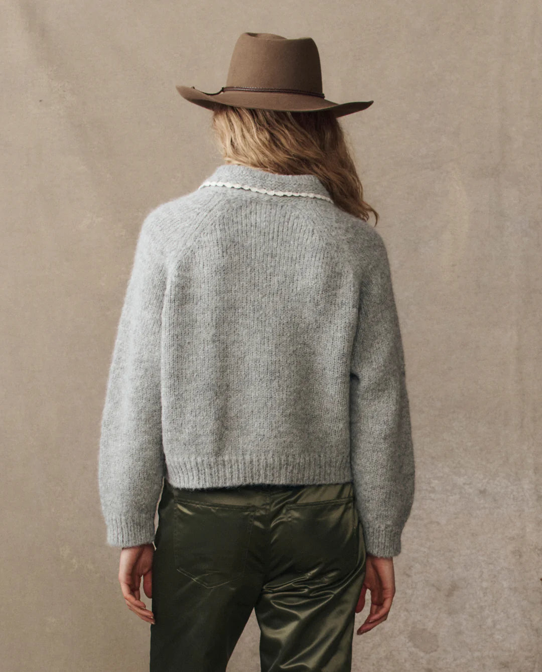 A person with wavy blond hair, wearing The Great Inc. Crochet Collar Pullover and shiny olive green pants, stands facing away from the camera against a beige background.