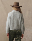 A person with wavy blond hair, wearing The Great Inc. Crochet Collar Pullover and shiny olive green pants, stands facing away from the camera against a beige background.