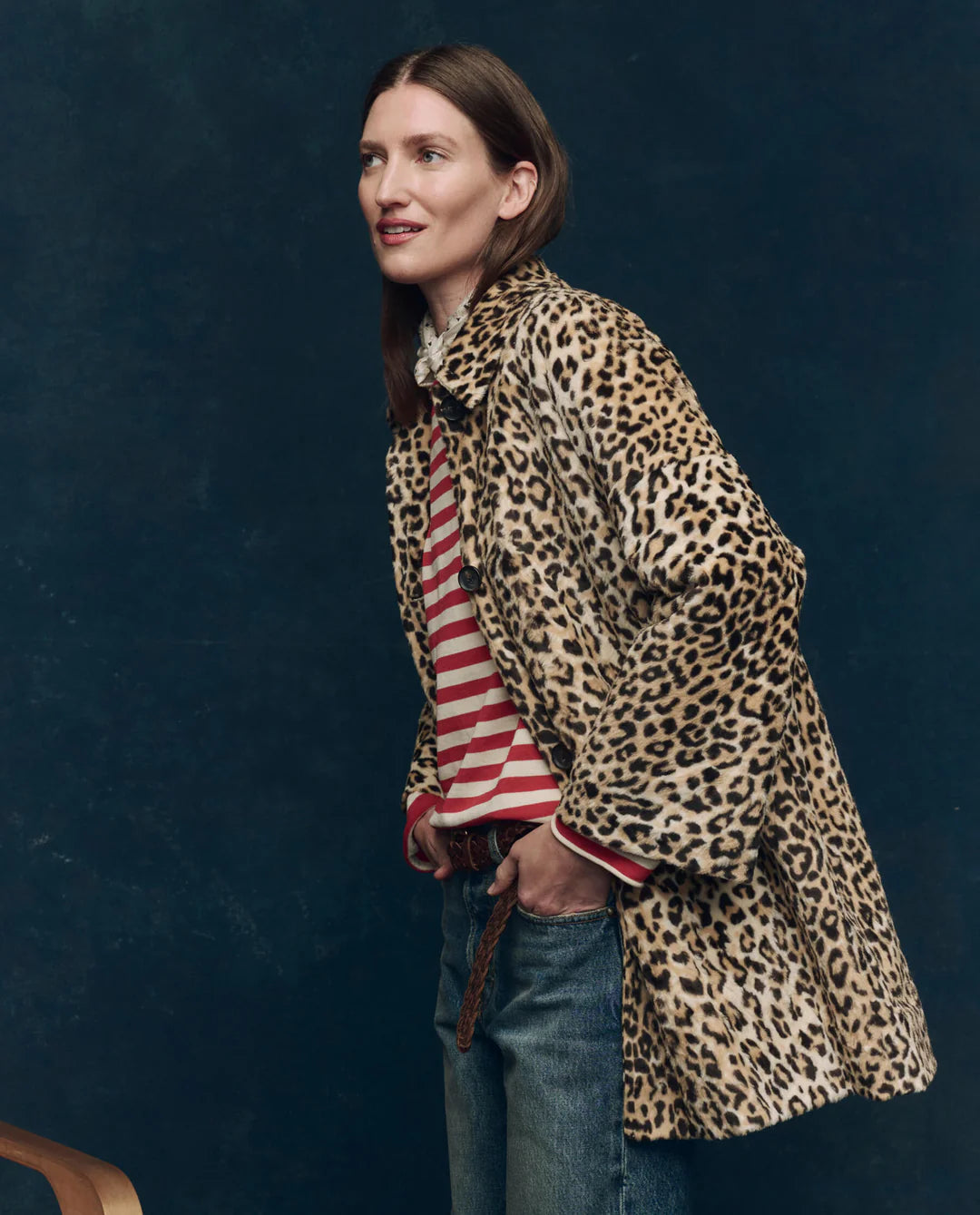 A woman wears The Car Coat by The Great Inc.—a crushed faux fur leopard print midcentury jacket—over a red and white striped shirt and blue jeans, standing against a dark textured background and smiling slightly.