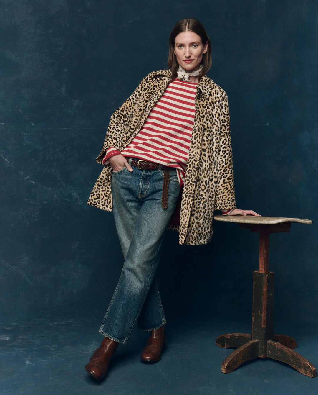 A person stands against a dark blue backdrop, wearing The Car Coat by The Great Inc.—a crushed faux fur leopard print midcentury design with quilted satin lining—over a red and white striped shirt, blue jeans, and brown boots, leaning on a rustic wooden table.