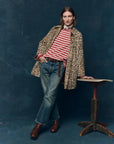A person stands against a dark blue backdrop, wearing The Car Coat by The Great Inc.—a crushed faux fur leopard print midcentury design with quilted satin lining—over a red and white striped shirt, blue jeans, and brown boots, leaning on a rustic wooden table.