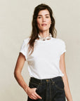 A woman with long brown hair models The Little Tee by The Great Inc., paired with dark high-waisted jeans and a chunky white necklace, standing against a plain beige background.
