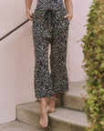 A person stands barefoot on concrete steps, wearing The Reef Pant by The Great Inc. in leopard-print cotton gauze with hands in pockets. A bush and pale pink wall in the background evoke relaxed seaside strolls.