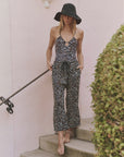 A woman in The Great Inc.’s The Reef Pant—leopard-print, sleeveless, with an elastic waistband—and a black wide-brim hat stands barefoot on steps against a pink wall and green shrub.