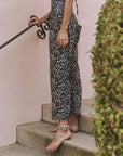 A barefoot person in The Great Inc. The Reef Pant with a leopard print and a shell anklet stands on concrete steps by a pink wall and leafy shrub, holding a black decorative handrail—ideal for carefree seaside strolls.