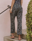 A barefoot person wears The Great Inc. Reef Pant in leopard print while standing on outdoor steps, holding a black railing, with greenery visible—a stylish choice for seaside strolls or relaxing afternoons.