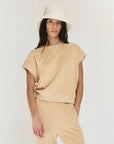 A woman with long dark hair wears The Micro Terry Wedge SS by The Great Inc., a beige short-sleeve top with matching pants, styled with vintage knits and a cream bucket hat, standing against a plain white background.