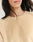 A woman with long dark hair wears The Micro Terry Wedge SS by The Great Inc., a beige sweatshirt with visible seam details. The photo captures only her lower face and upper torso, emphasizing its subtle vintage-inspired style.