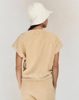 A person faces away from the camera, wearing The Great Inc.'s The Micro Terry Wedge SS in beige with matching pants and a white bucket hat, set against a plain light background.