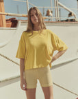 A woman stands outdoors by a boat, wearing The Micro Terry Track Crew by The Great Inc., a loose yellow T-shirt with an oversized silhouette, and yellow striped shorts. Her long light brown hair catches the sun as she looks at the camera on a bright day.