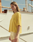 A woman with long hair stands outdoors by a white boat under a clear sky, wearing The Great Inc.’s Micro Terry Track Crew—a loose yellow cotton top—paired with light-colored shorts.