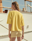 A woman with light brown hair stands on a boat, facing away, wearing The Micro Terry Track Crew by The Great Inc. in yellow and light beige shorts. The sunny sky and parts of the boat’s railing are visible.