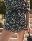 A person wears The Delta Short by The Great Inc.—a long-sleeved, button-up romper with a black, brown, and white leopard print and an elastic waistband—standing outdoors and holding a small black textured bag.