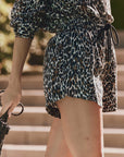 A person stands outdoors wearing The Great Inc.'s The Delta Short with an elastic waistband and a matching leopard print top, holding a black textured handbag. Blurred stone steps and greenery appear in the background.
