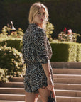 A woman with long blonde hair stands on stone steps outdoors, wearing The Delta Short by The Great Inc., featuring a leopard print and elastic waistband, while looking over her shoulder amid lush greenery and flowers.