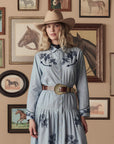 A woman in a tan cowboy hat and blue dress with dark blue embroidery stands before framed horse paintings. She has wavy blonde hair and wears The Great Inc.’s The Big Buckle Belt, featuring Western craftsmanship and a large buckle.