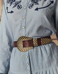 A person wearing a light blue button-up dress with dark blue floral embroidery and The Big Buckle Belt by The Great Inc., made of top-grain leather and featuring a wide design with a gold sculpted buckle and tip.