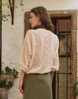 A woman with loose brown waves stands near a rustic stone wall and window, wearing The Great Inc.'s The Crochet Pullover and a green skirt.