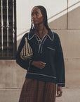 The Great Inc. menswear polo with white trim and a plaid skirt, styled outdoors with a striped bag.