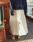 A person stands on a wooden boat deck with their back to the camera, wearing The Viola Skirt by The Great Inc., paired with a navy patterned sweater and tan slip-on shoes.