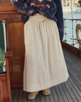 Wearing The Great Inc.'s Viola Skirt—an ankle-length cream cotton skirt with a smocked waistband—a person stands on a wooden boat deck, paired with tan shoes and a navy star-patterned sweater; water and buildings are visible behind.