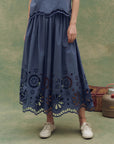Wearing The Great Inc. Super Bloom Eyelet Skirt with floral eyelet cutouts at the hem, a person stands on a mottled green surface in white shoes, with a vintage picnic basket and metal container behind them.