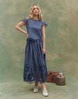 A woman wears The Great Inc. Super Bloom Eyelet Skirt, featuring floral eyelet cutouts, paired with white sneakers. She stands by a vintage basket and metal canister against a green textured backdrop.