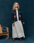 A woman with curly blond hair wears a long white dress, brown boots, and The Great Inc.'s The Horse Lodge Cardigan, featuring hand-embroidered horses, standing by a wooden chair against a dark blue backdrop for western nostalgia.