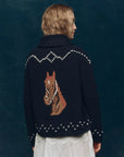 A person with wavy blond hair wears The Great Inc.'s Horse Lodge Cardigan—a dark blue knit sweater with a large hand-embroidered horse and white stitching—paired with a white skirt, standing against a dark blue background.