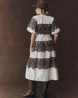 A person wearing The Fairground Dress by The Great Inc., a vintage cotton dress with brown lace patterns, and cowboy boots stands facing away against a brown backdrop.
