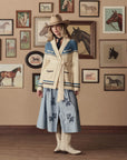 A woman in The Great Inc.'s The Western Riding Cardigan with blue accents, a blue floral skirt, and white cowboy boots stands indoors by horse paintings, perfectly embodying classic Western Americana style.