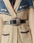 A person wearing a thick, cream cardigan with blue patterns and dark trim, cinched at the waist by The Great Inc.'s Big Buckle Belt—crafted from top-grain leather and featuring a large decorative silver buckle inspired by Western style.