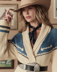 A woman in a beige cowboy hat, black bandana, and silver-buckled belt poses indoors in The Western Riding Cardigan by The Great Inc.—a patterned sweater with blue accents—lifting her hat’s brim in classic Western style before framed pictures.
