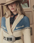 A woman in a cowboy hat and The Big Buckle Belt by The Great Inc. wears a cream and blue cardigan with western patterns and a dark bandana, standing before framed pictures on a beige wall.