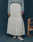 A person wearing a blue-and-white striped long-sleeve shirt, The Great Inc.'s The Victorian Skirt, and white high-top sneakers stands next to a wooden chair against a dark blue background.