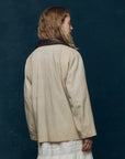 A person with wavy, light brown hair is seen from behind wearing The Great Inc.’s cream-colored Barn Jacket with a dark collar over a white dress, standing against a dark teal background.