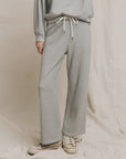 A person wears The Great Inc.'s The Lap Sweatpant and matching sweatshirt in light gray, paired with white sneakers, standing on a beige fabric backdrop. Only their body from the shoulders down is shown. This set is made in Los Angeles.