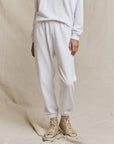 Dressed in a white long sleeve shirt and The Stadium Sweatpant by The Great Inc., the person stands against a neutral backdrop wearing beige high-tops with white laces, conveying a vintage athletic style. The image centers on their lower body.