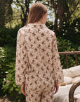 Seen from behind, a person with long hair stands outdoors in The Pajama Shirt by The Great Inc.—a beige top with a brown and green horse pattern. Lush foliage and a metal bed frame add to the vintage sleepwear vibe.